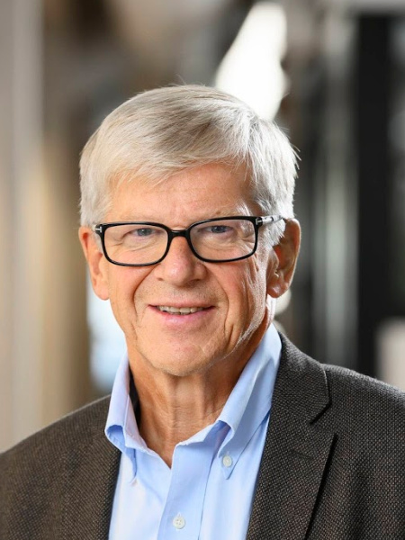 En man med grått hår och svarta glasögon, blå skjorta och en mörkbrun kavaj ler mot kameran