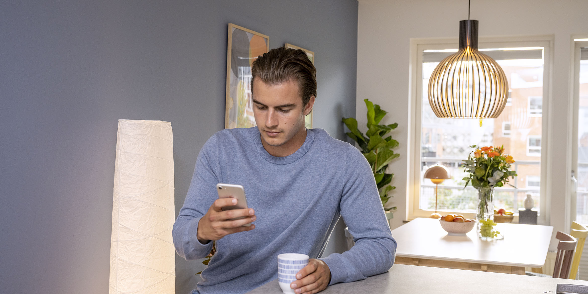 En kille i blå tröja sitter vid en köksö och dricker kaffe och kollar på sin mobiltelefon