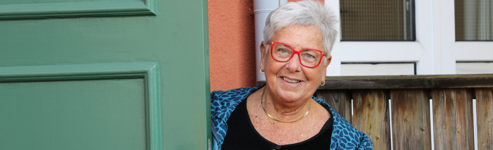 En gråhårig kvinna med röda glasögon står vid en grön dörr