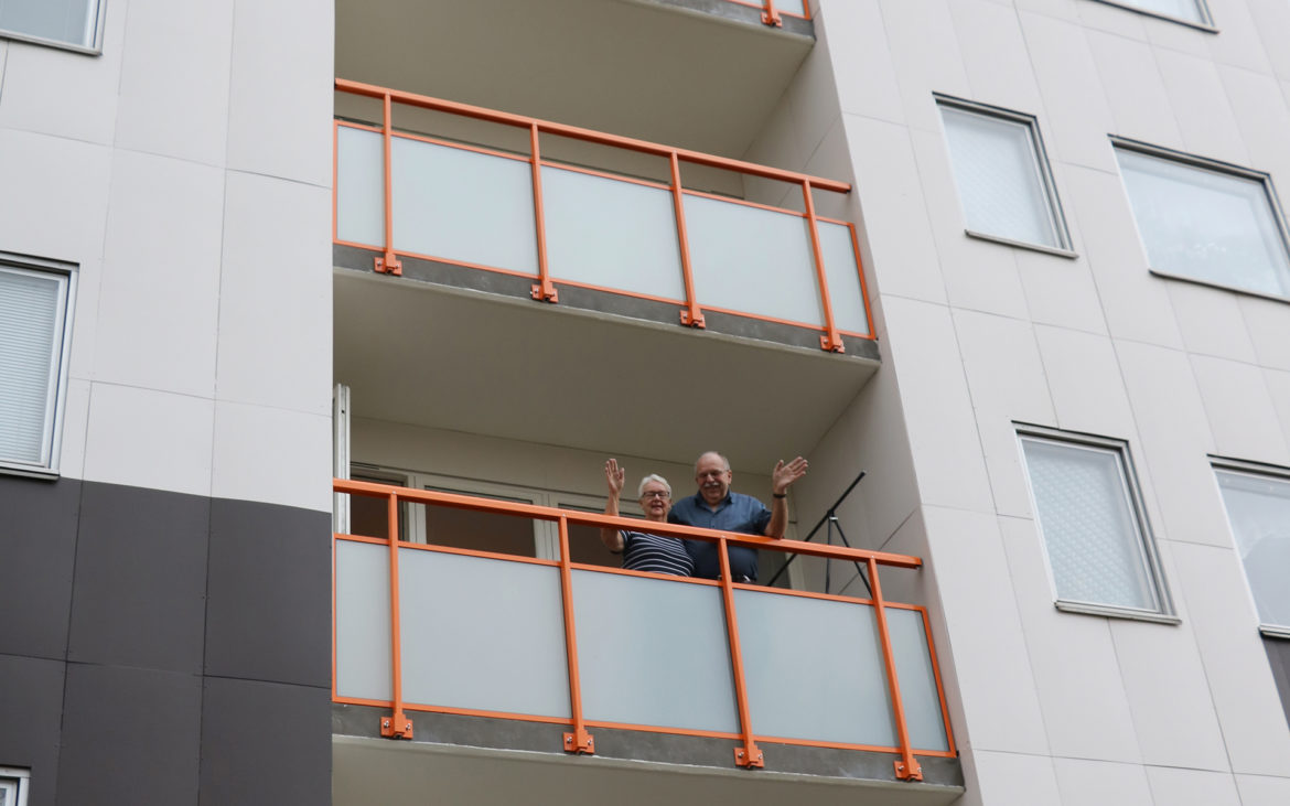 Ett bostadshus där två personer står på en balkong och vinkar ner på gatan