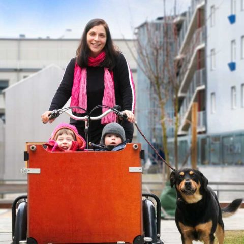 En kvinna i svart jacka och rosa halsduk som cyklar på en lådcykel med två barn som sitter i. Bredvid springer en hund som är kopplad.