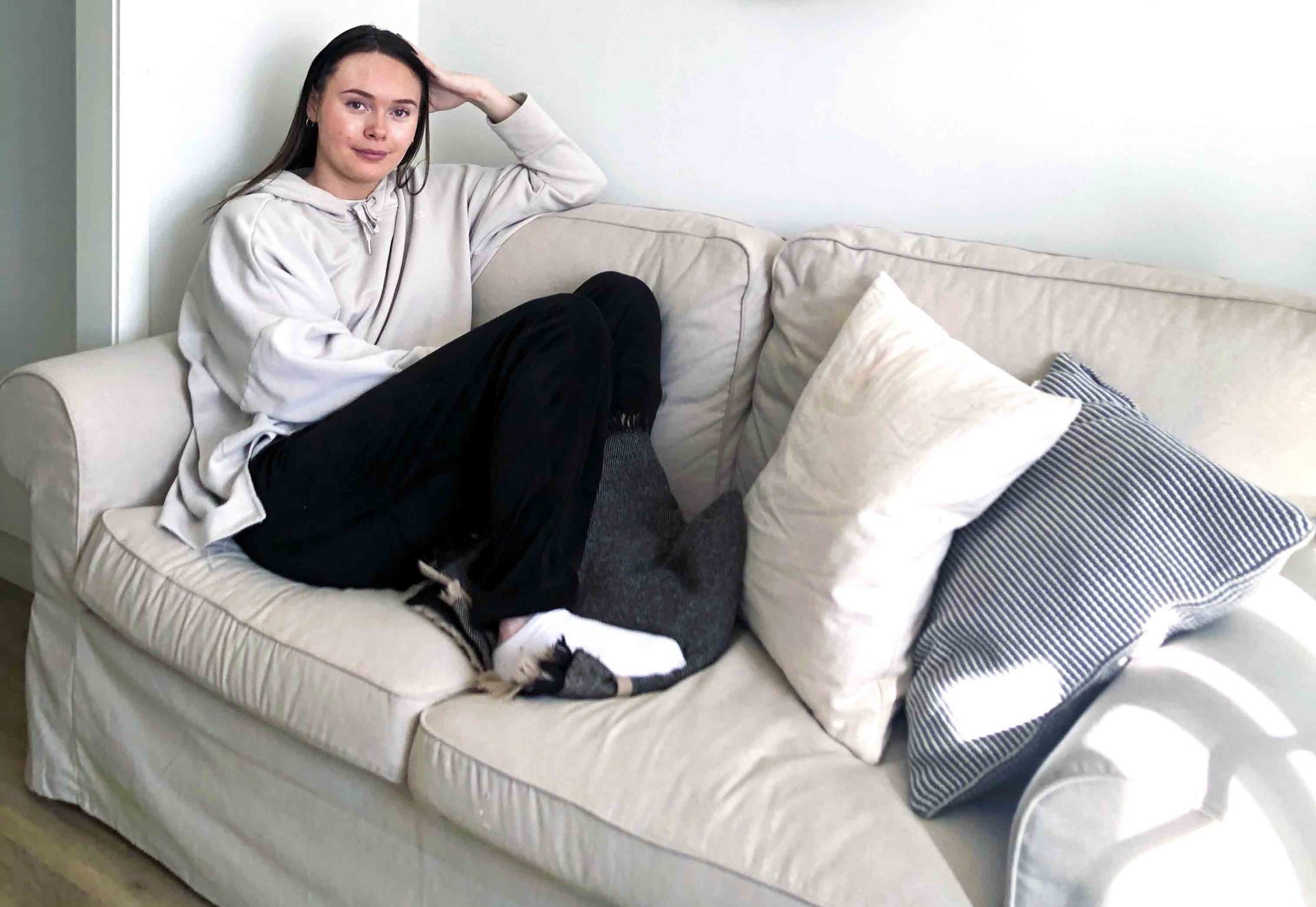 En tjej med mörkt hår och en grå tröja sitter i en beige soffa med massa kuddar