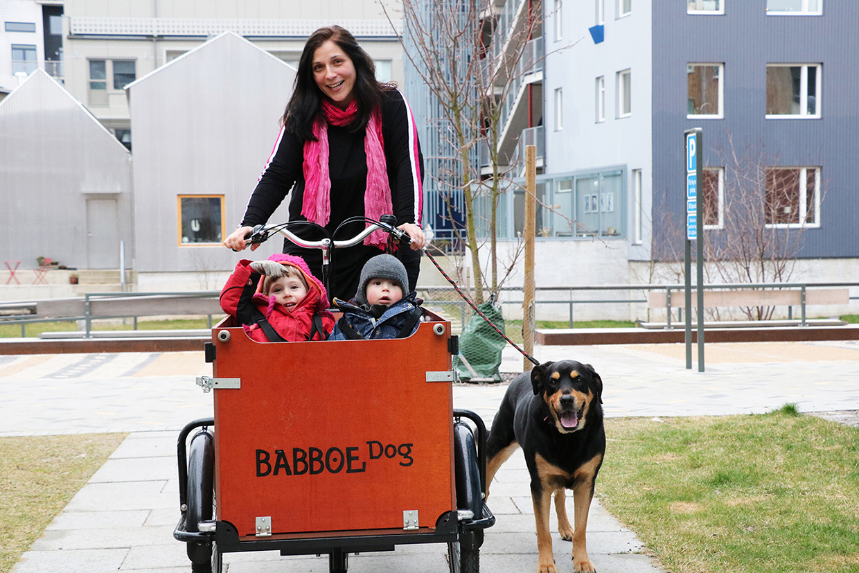 En kvinna med en rosa halsduk cyklar på sin lådcykel med en hund bredvid