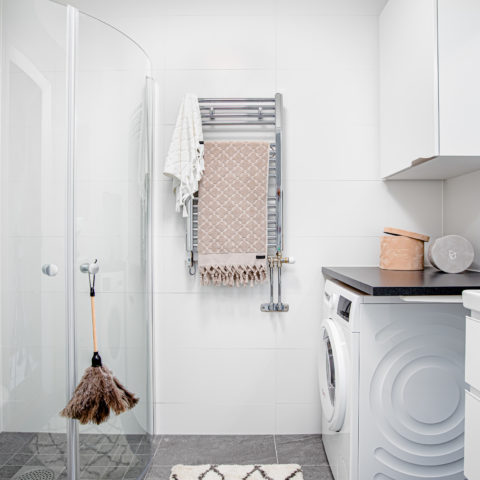 Ett ljust badrum där man ser en duschvägg i glas, handdukstork i krom och en vit tvättmaskin.