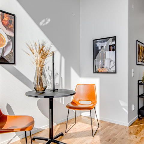 Matdel i ett kök där två orange platsstolar och ett svart litet runt bord står. På väggen hänger det två tavlor med motiv av kaffebönor och koppar.