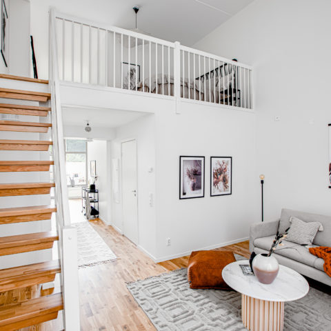 Ett ljust rum där man ser en beige soffa med orange filt i. Bakom ser man en trappa i trä som går upp till ett sovloft.