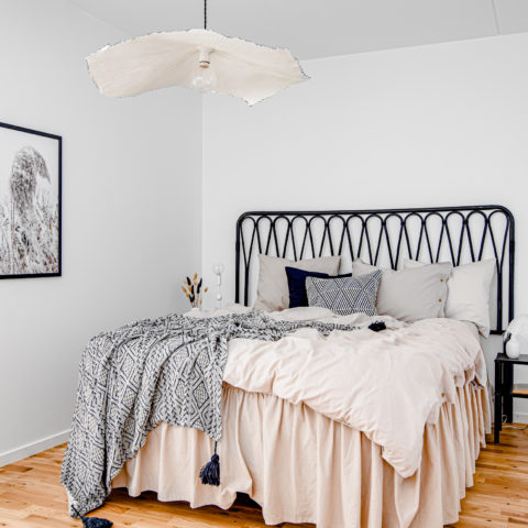 En säng med en stor rosa sängkappa och massa kuddar. I taket hänger en stor beige lampa.