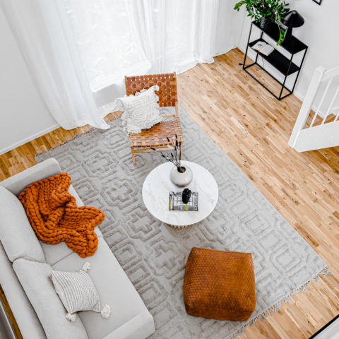 Foto taget uppifrån och ner i ett vardagsrum. En mönstrad beige matta med en vit soffa där orange inredningsdetaljer är med