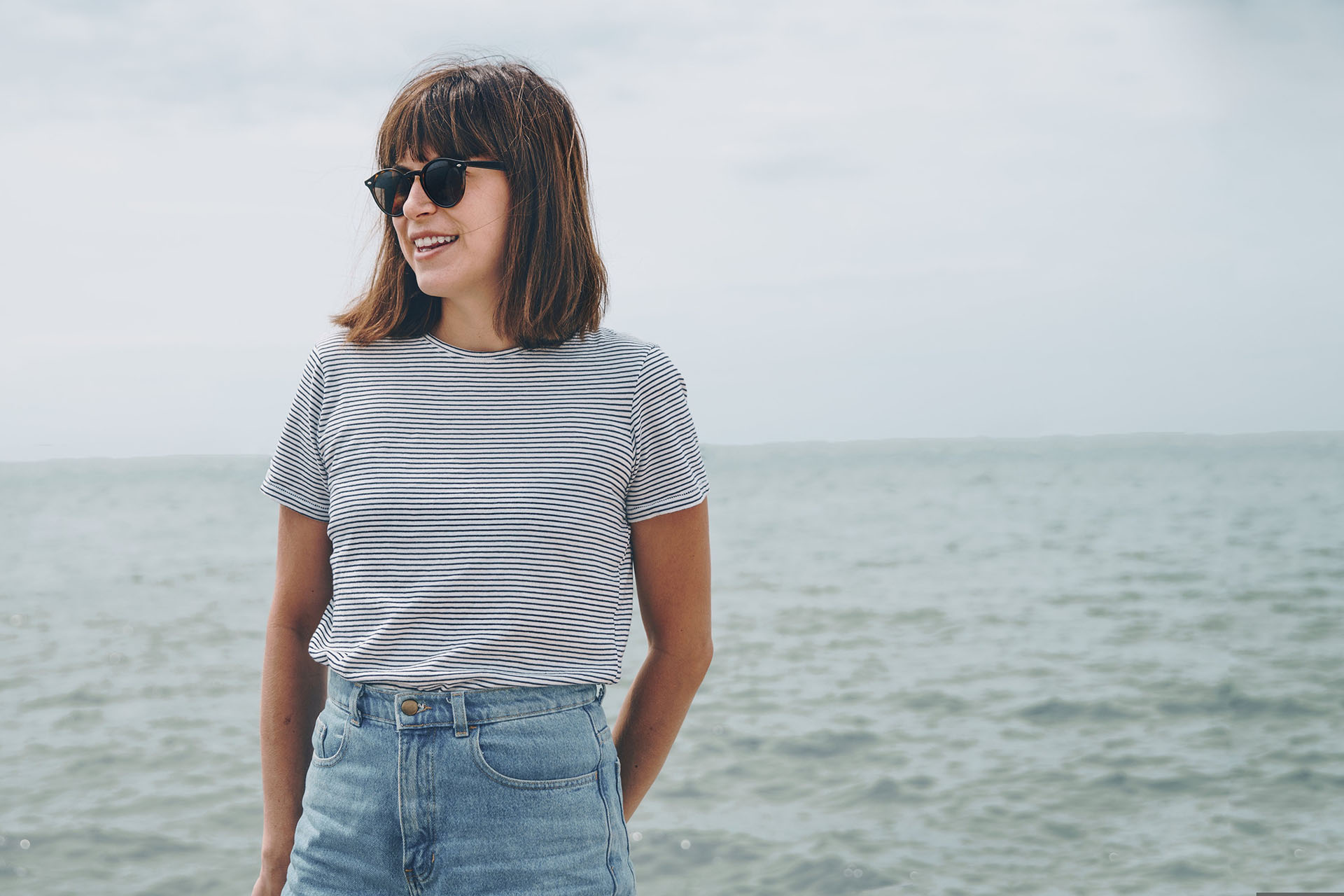 En brunhårig tjej med randig tröja står vid havet i solglasögon