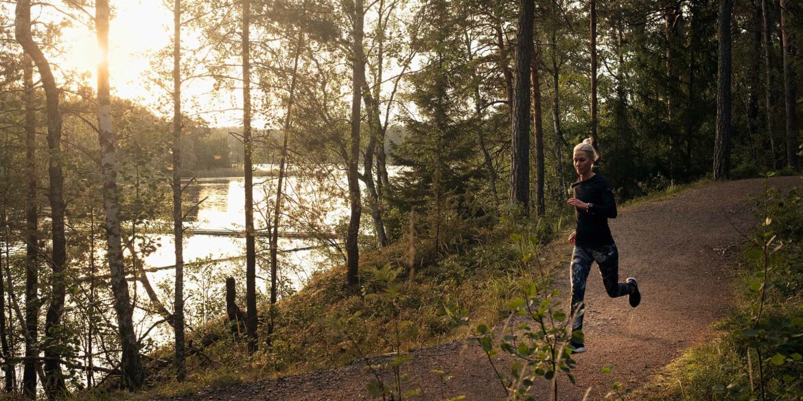 En ljushårig tjej springer i en skog där man ser en sjö i bakgrunden och där solen är på väg ner