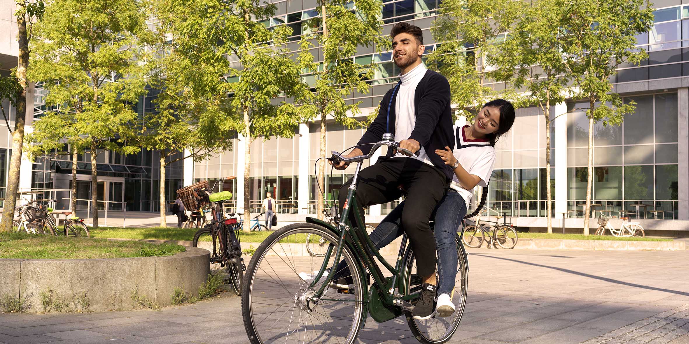 En kille med mörka kläder cyklar på en cykel med en tjej sittandes på pakethållaren