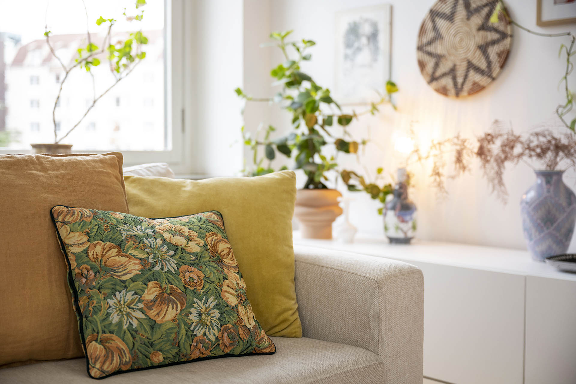 En ljus beige soffa med en blommig och gula kuddar. Bakom syns en grön krukväxt och en lampa som lyser