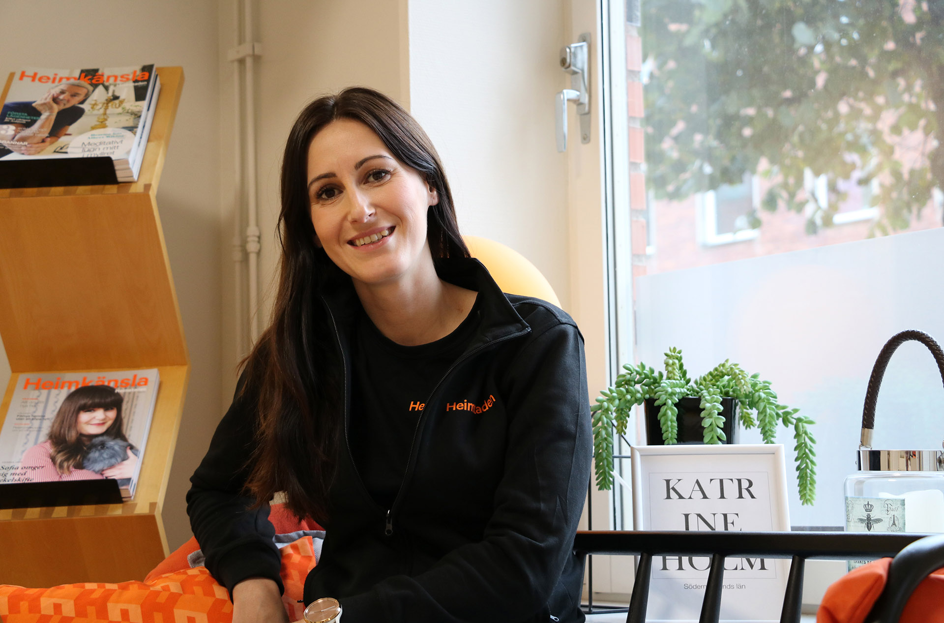 En kvinna i mörkt långt hår sitter bredvid en tidningshylla och tittar in i kameran