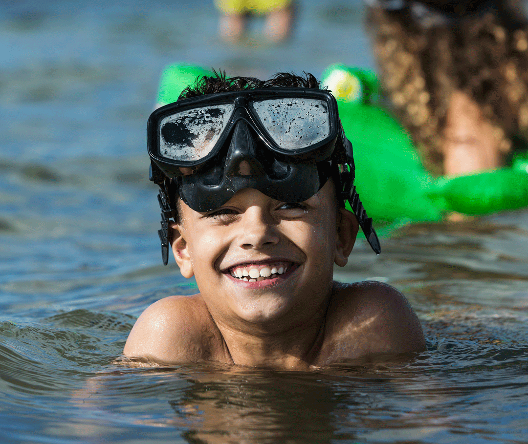 En pojke som badar i havet med ett par snorkelglasögon på huvudet. Bakom honom ser man en tjej som har en grön badring.