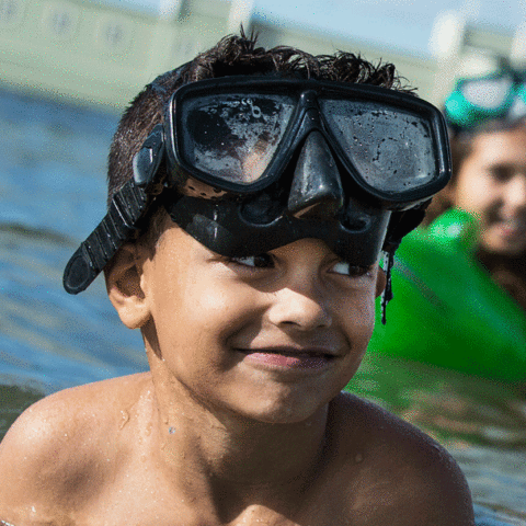En pojke som badar i havet med ett par snorkelglasögon på huvudet. Bakom honom ser man en tjej som har en grön badring