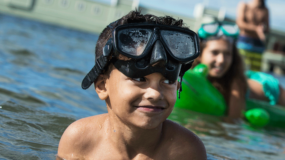En pojke som badar i havet med ett par snorkelglasögon på huvudet. Bakom honom ser man en tjej som har en grön badring