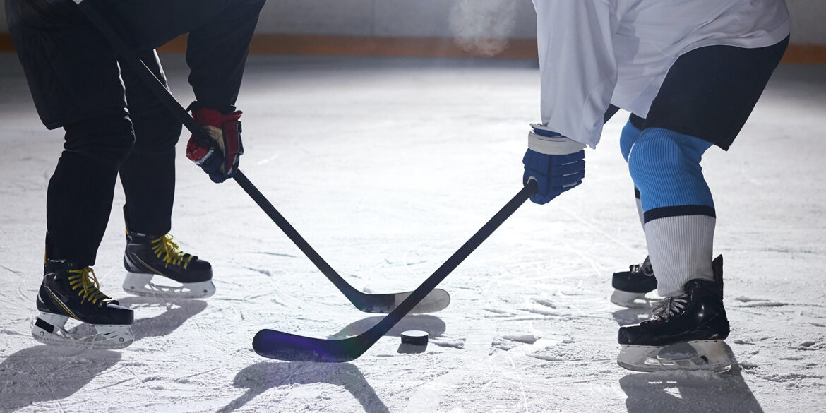 Två personen som är klädda i hockeyutrustning står på en isig bana med klubborna mot varandra.