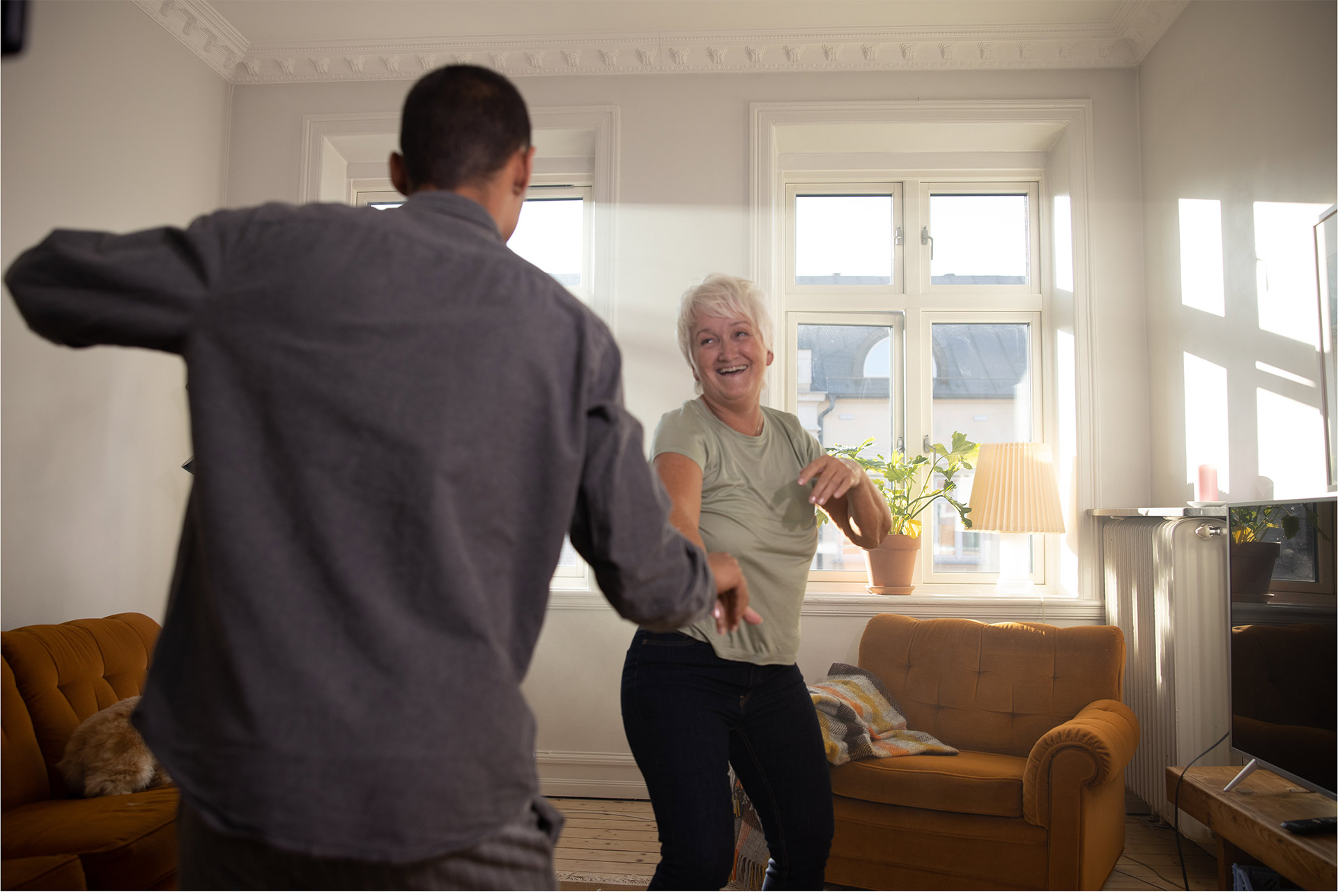 En äldre kvinna och en yngre man dansar i vardagsrummet. Bakom de ser man en orange soffa och en gulorange fåtölj.