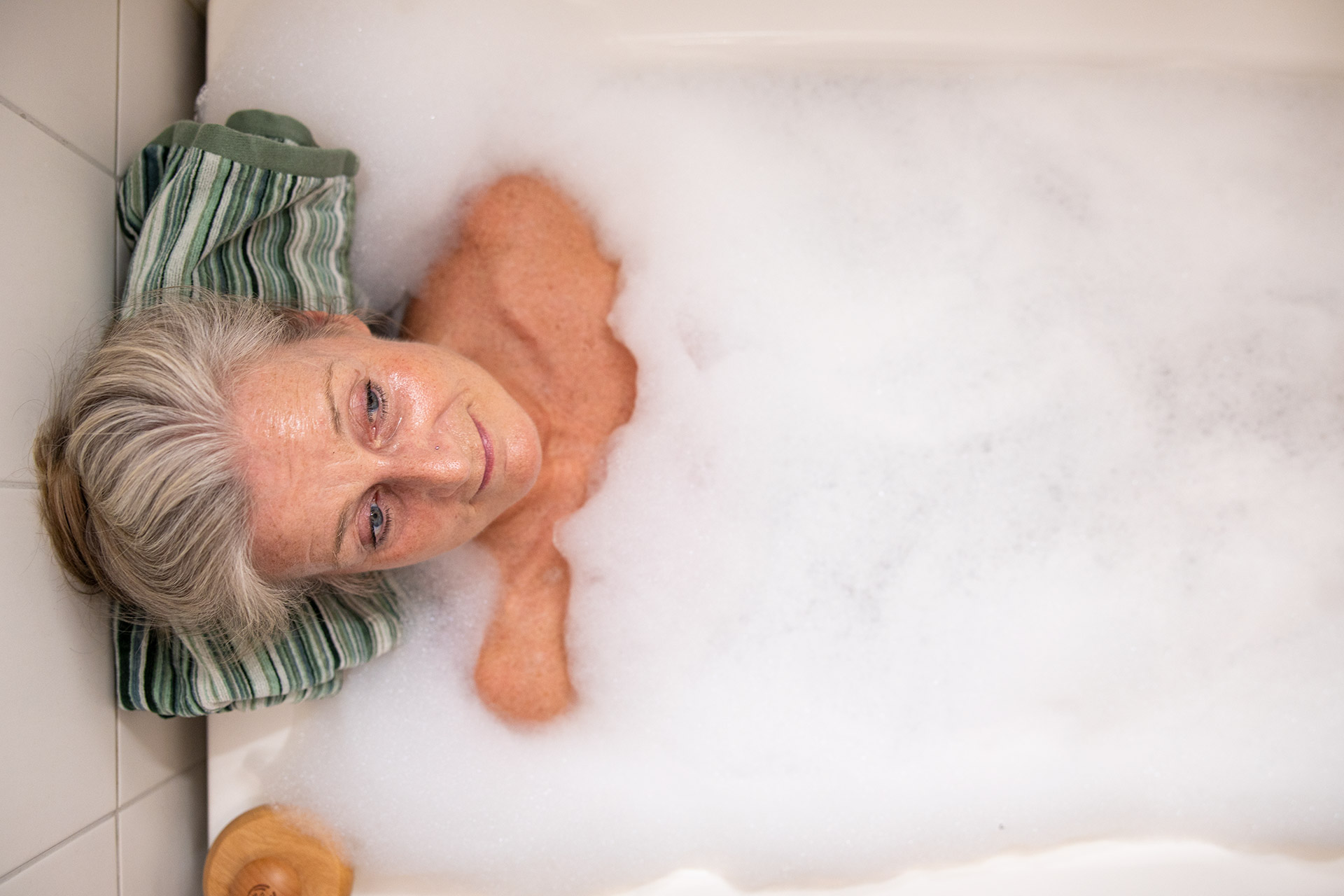 En kvinna med grått hår ligger i ett badkar som är fyllt med badskum. Under hennes nacke har hon en randig handduk.