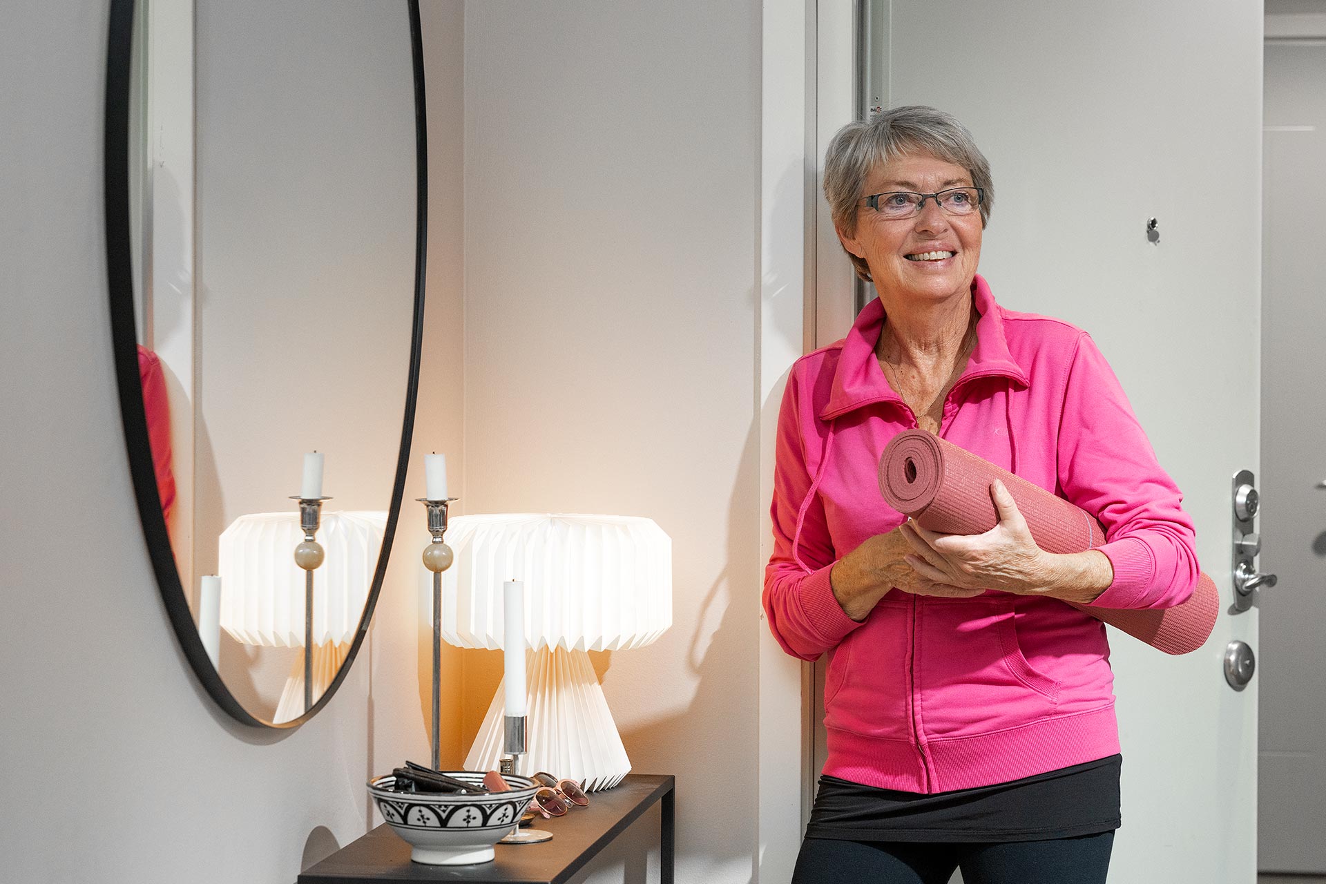 En gråhårig äldre kvinna står lutad mot en vägg med en rosa yogamatta under armen. I rummet hänger en stor oval spegel och på byrån står det en lampa och lyser.