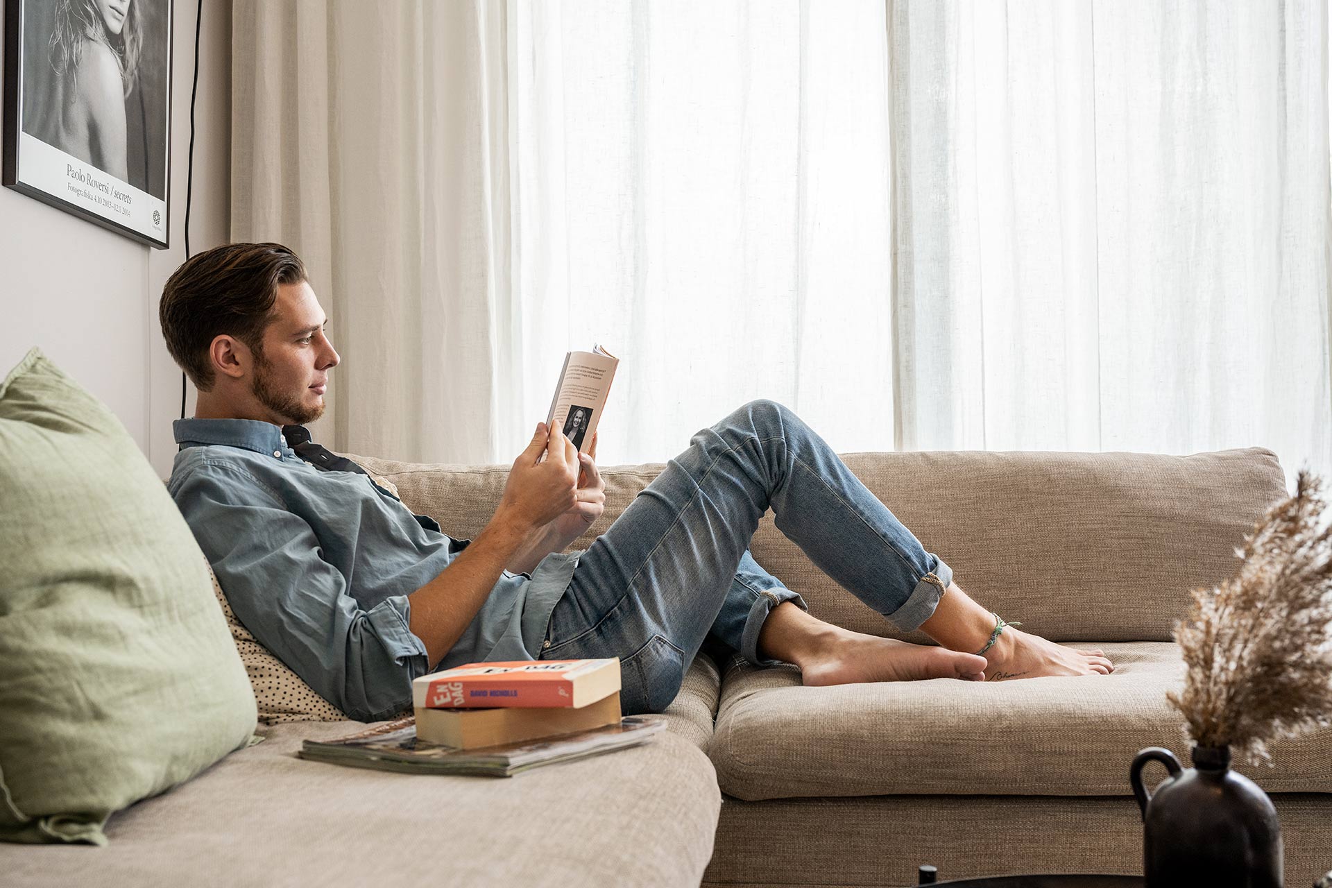 En man iklädd jeans och jeansskjorta ligger i sin soffa och läser en bok. I soffan ligger det fler böcker.
