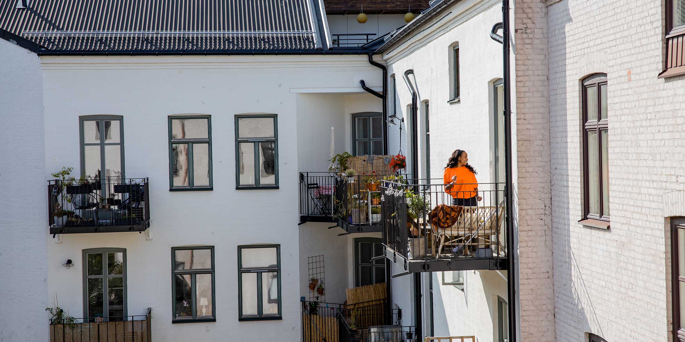 Ett vitt putsat hyreshus där man ser balkonger med gröna växter på. På en av balkongerna står en kvinna med mörkt hår och en orange tröja och kollar in i lägenheten.