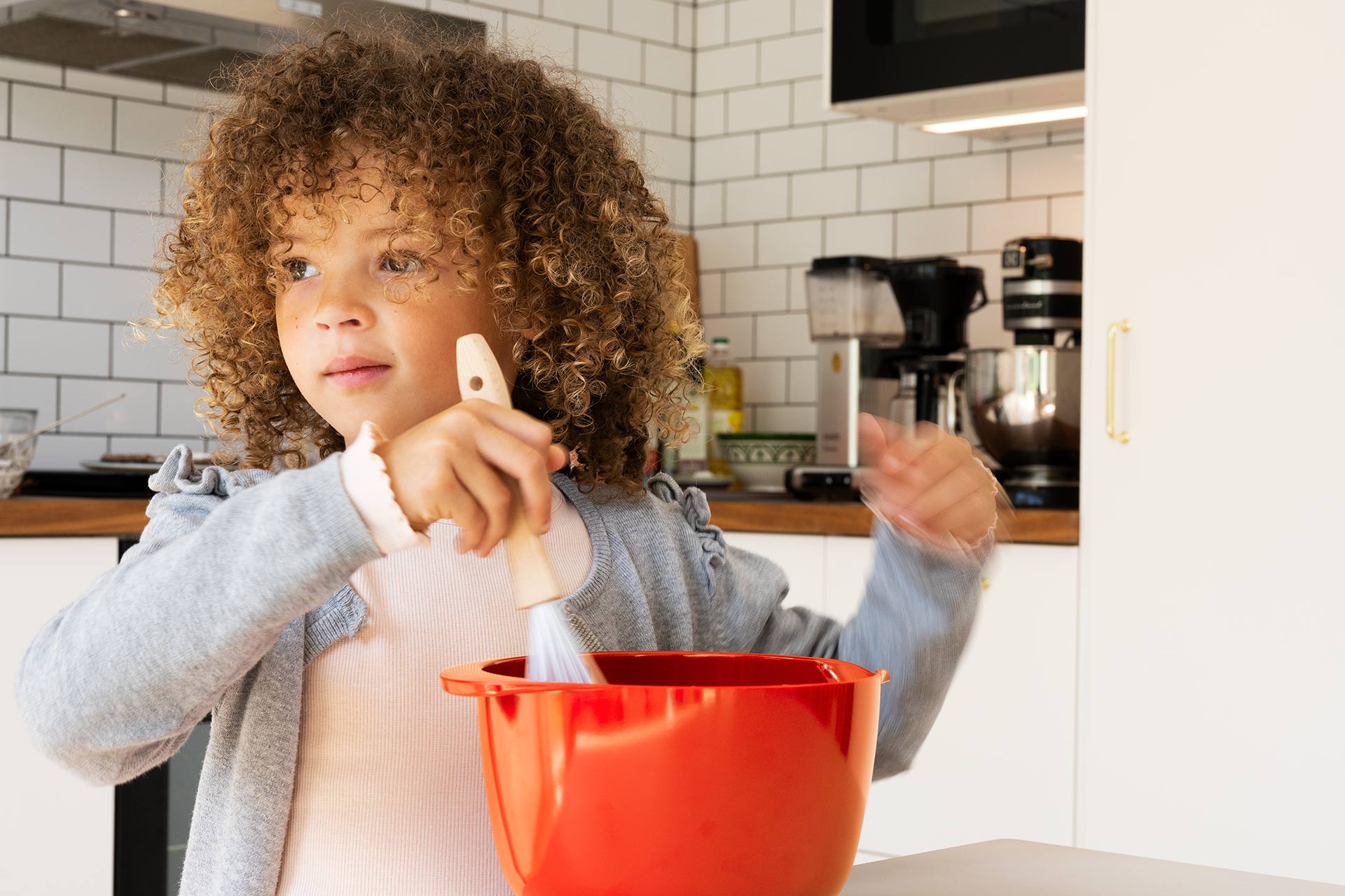 Ett barn med mörkt lockigt hår vispar i en röd skål i köket