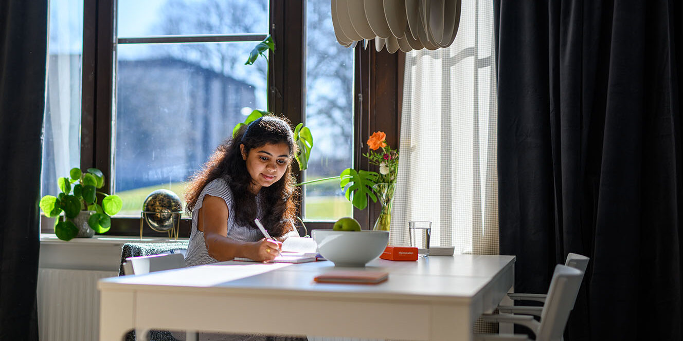 En ung mörkhårig tjej sitter vid sitt köksbord och skriver i ett block. Bakom henne ser man stora fönster och gardiner.