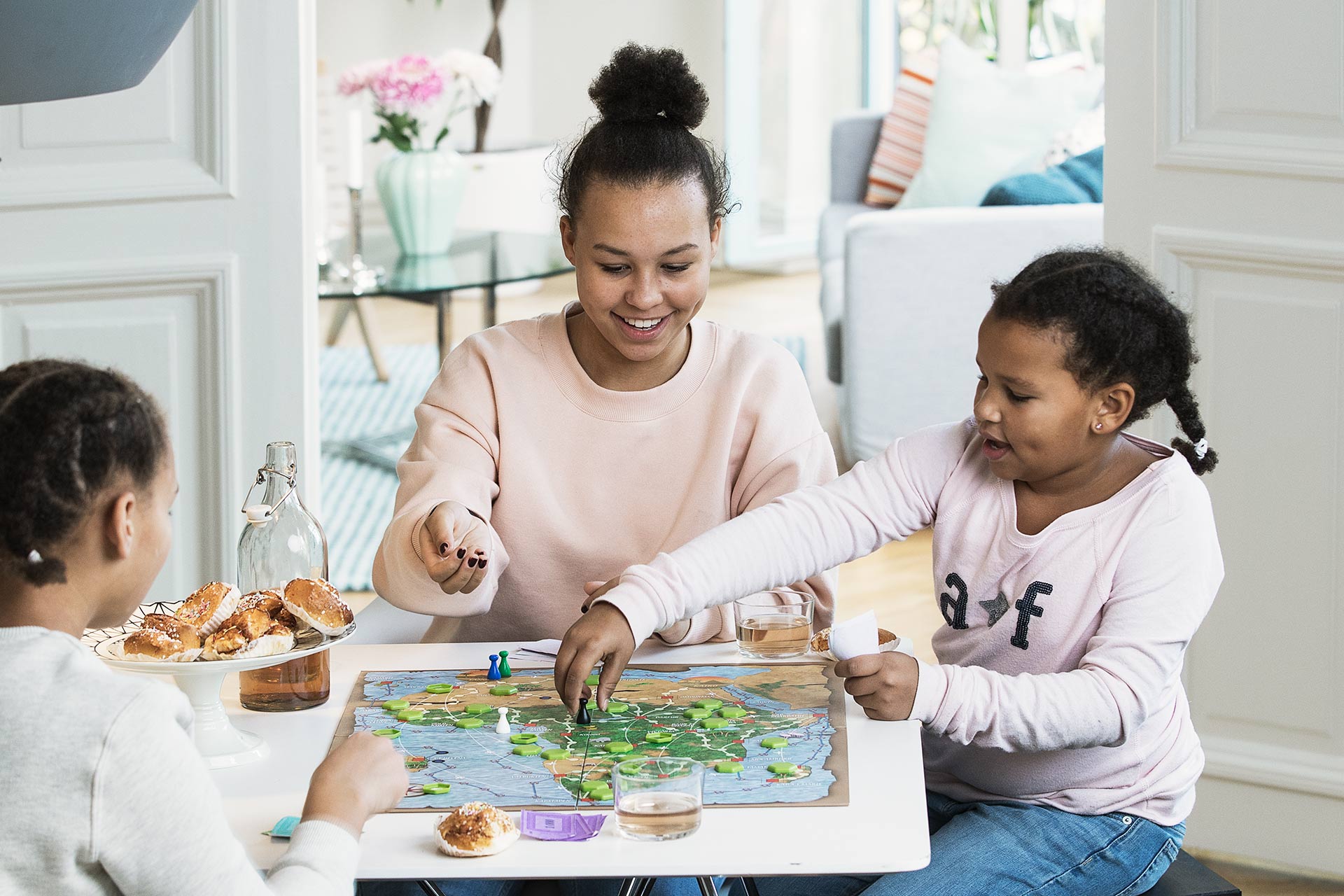En kvinna i rosa tröja och två barn sitter vid ett bord i hemmet och spelar spel. På bordet står det ett fat med kanelbullar.