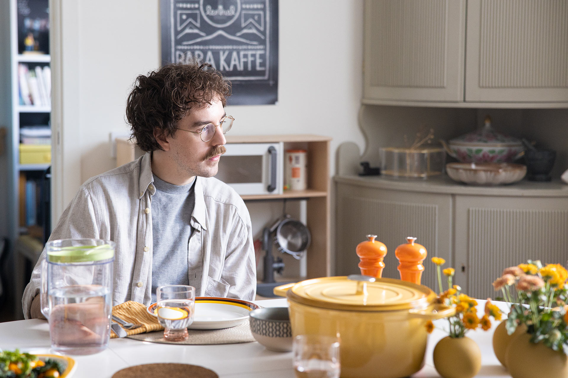 En man med lockigt mörkrött hår och en beige skjorta sitter vid köksbordet och tittar åt sidan. På bordet står det en gul kastrull och salt och pepparkvarn.