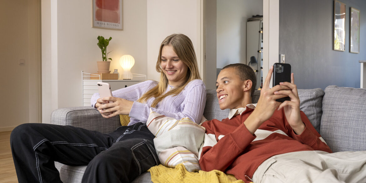 En ung tjej och kille halvligger i en grå soffa och tittar på sina mobiler och skrattar båda två.