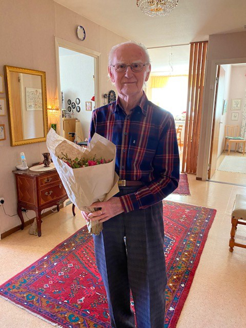 En äldre man står med en bukett blommor i famnen i sitt hem.