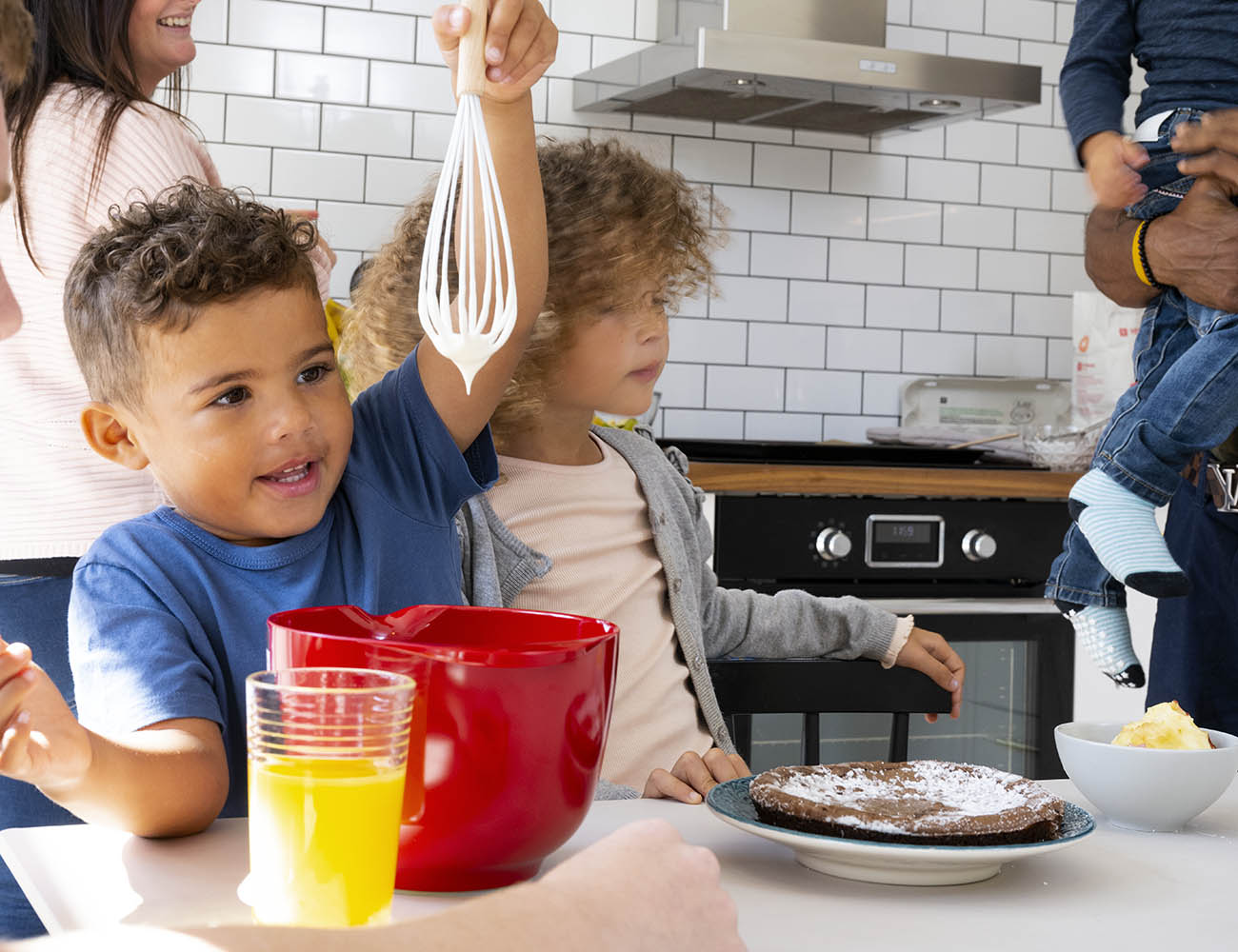En pojke med blå tröja står i ett kök och håller upp en visp som är full med vit smet från en röd bunke. Bredvid henne står en tjej med lockigt brunt hår som sneglar på ett fat med pannkakor