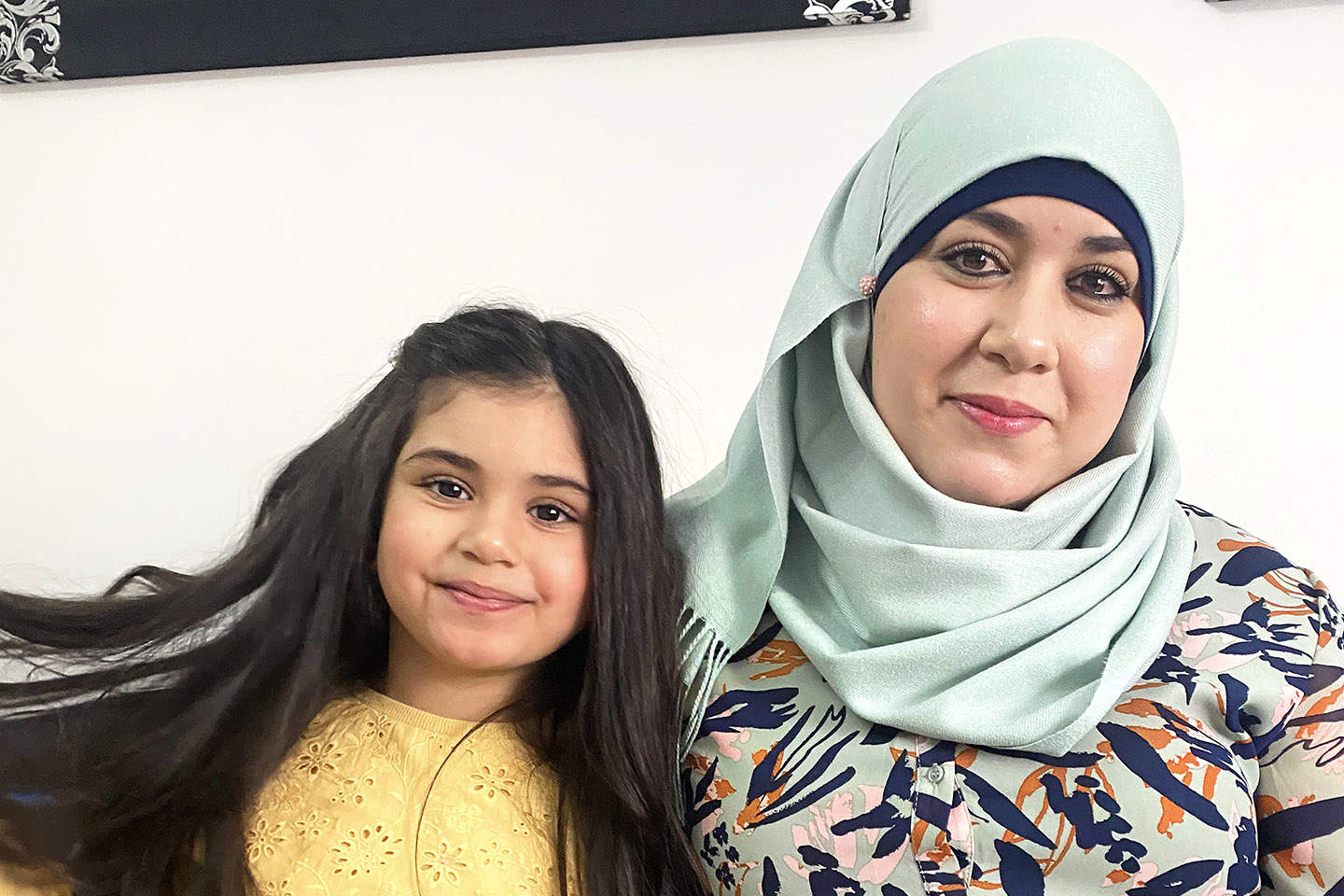 En kvinna och ett barn med mörkt långt hår, sitter i en soffa och ler mot kameran