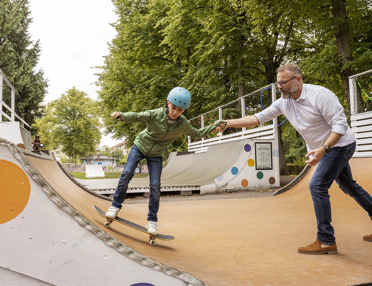 En man i skjorta och jeans hjälper en pojke i en skateboardramp som åker på en bräda med en blå hjälm.