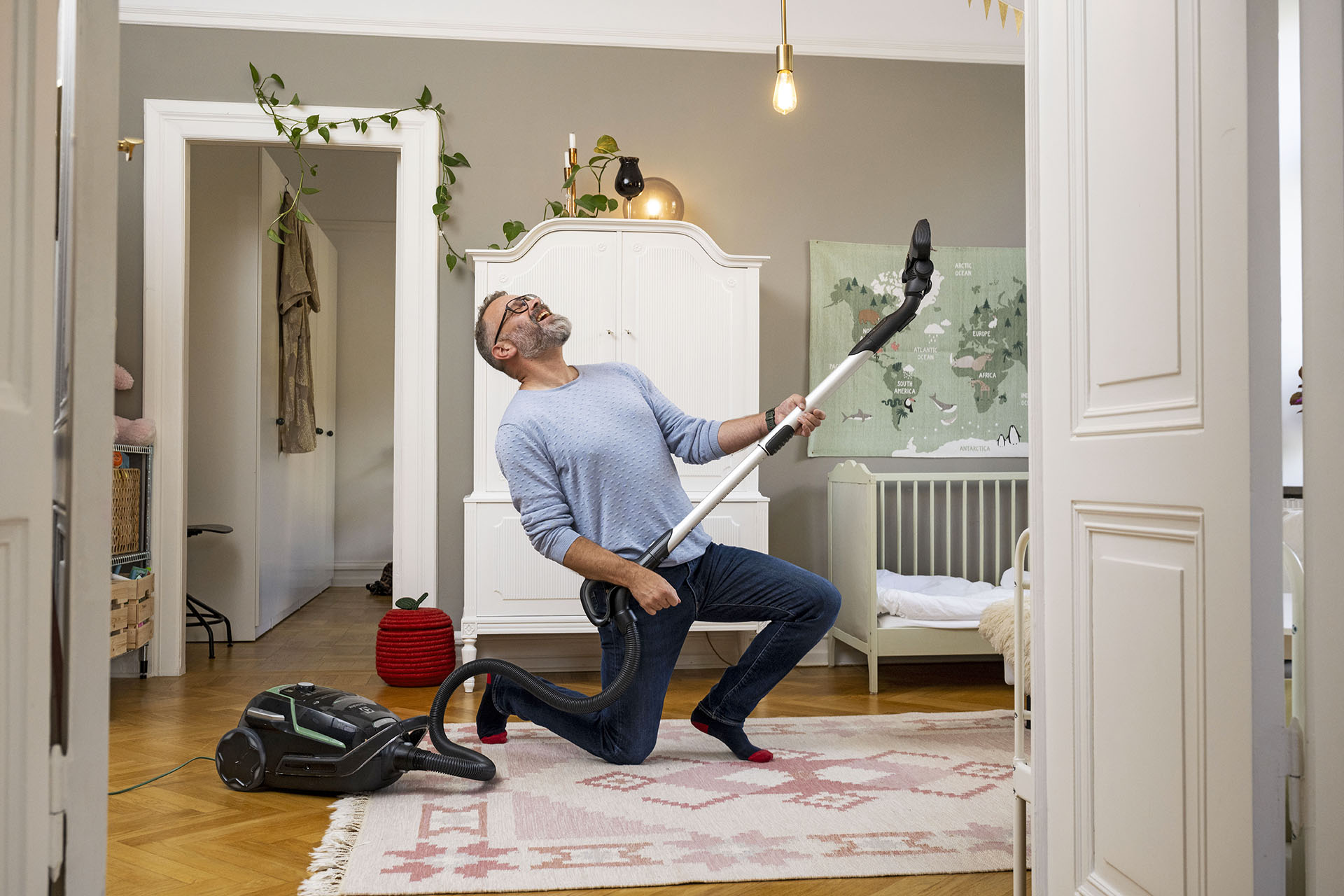 En man med blå tröja står i sitt vardagsrum och håller i sin dammsugare som om det är en gitarr. Bakom honom ser man ett stort vitt skåp med en lampa på.