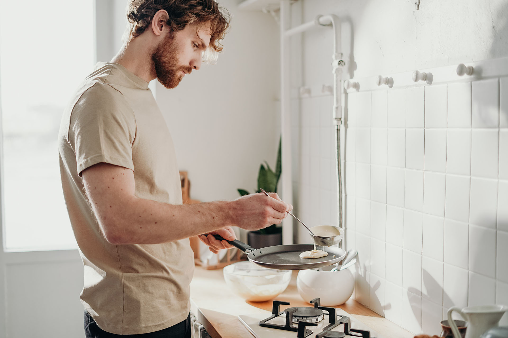 En kille i en beige kortärmad tröja står i ett kök med en stekpanna i handen