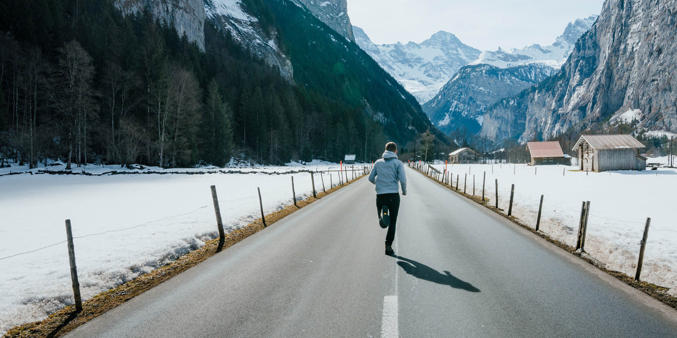 En kille i grå hoodtröja och svarta byxor springer på en ödslig väg där man ser höga berg, snö och några gamla hyddor på sidan.
