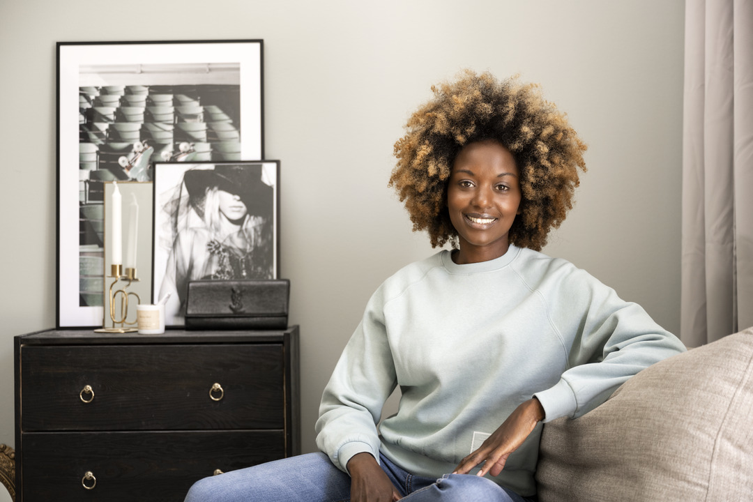 En tjej med stort lockigt hår sitter i en beige soffa och ler in i kameran. Bakom henne ser man en brun byrå med några tavlor ståendes på.