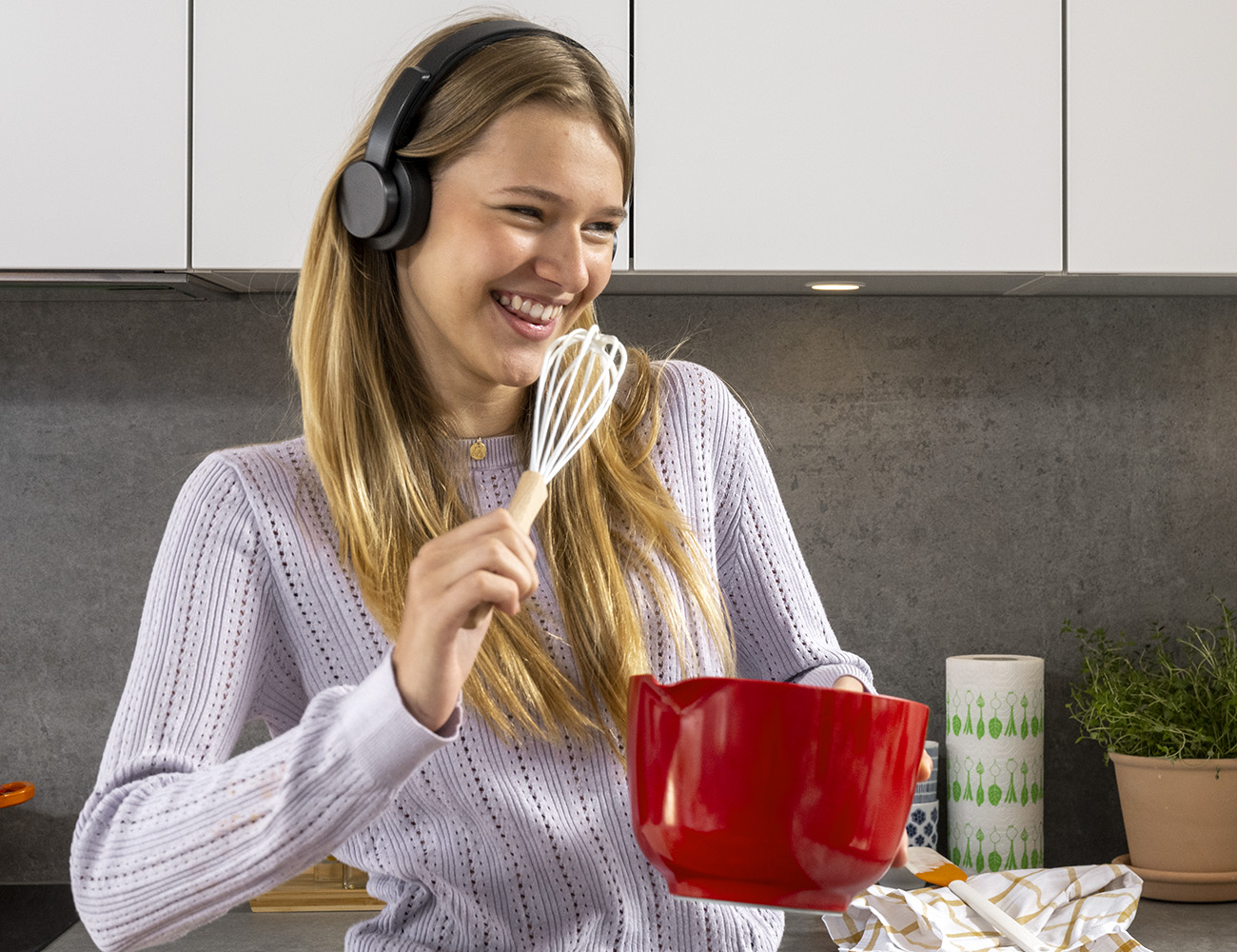 En ung tjej med långt hår och svarta hörlurar står i ett kök, med en röd plastbunke i handen och sjunger och låtsas att vispen är en mikrofon.