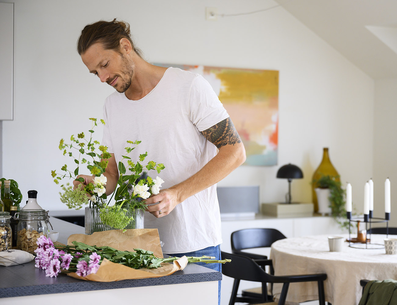En ung man i vit tshirt och tatueringar på armen står i sitt vardagsrum och sätter en blombukett i en glasvas