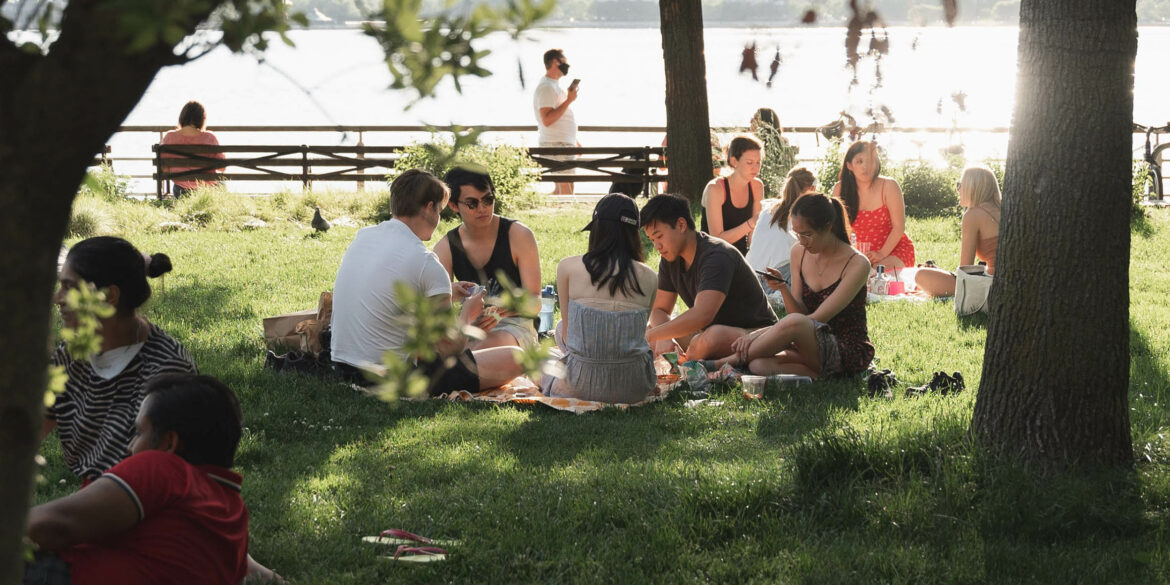 Massa sommarklädda människor sitter i en park vid vattnet. De sitter på filtar och dricker och äter.