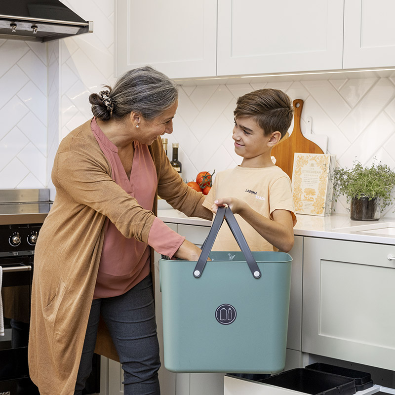 En kvinna med gråsvart hår och en ung pojke med gul tröja står i ett kök med en sopsorteringslåda i handen och ler mot varandra