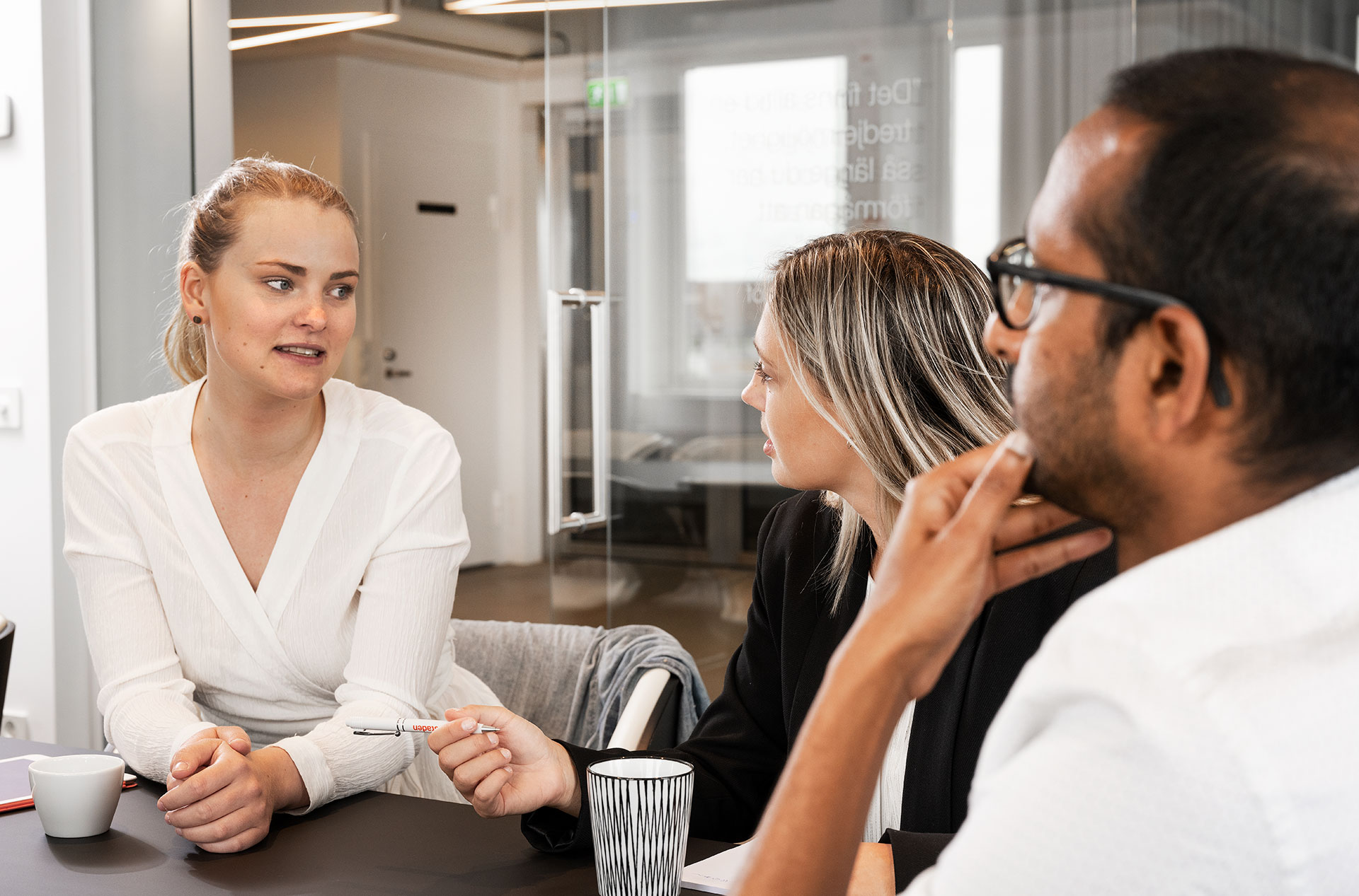 Två kvinnor och en man sitter vid ett svart bord i en kontorsmiljö och pratar med varandra. På bordet står det kaffemuggar i svart och vitt