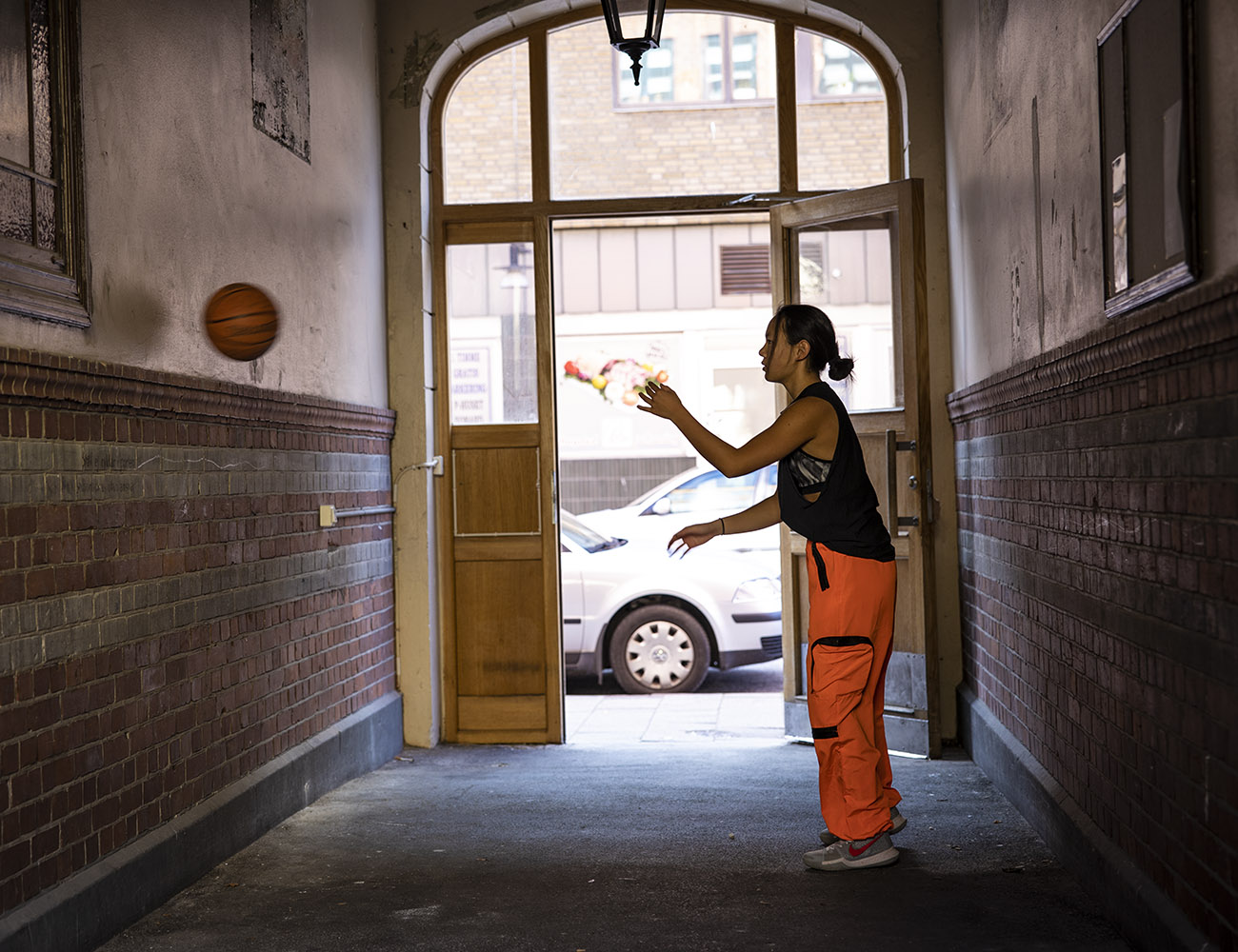 En tjej i orange byxor och svart linne står i trapphus entrén och kastar en basketboll mot väggen. Porten står öppen och utanför ser man en bil som står parkerad.