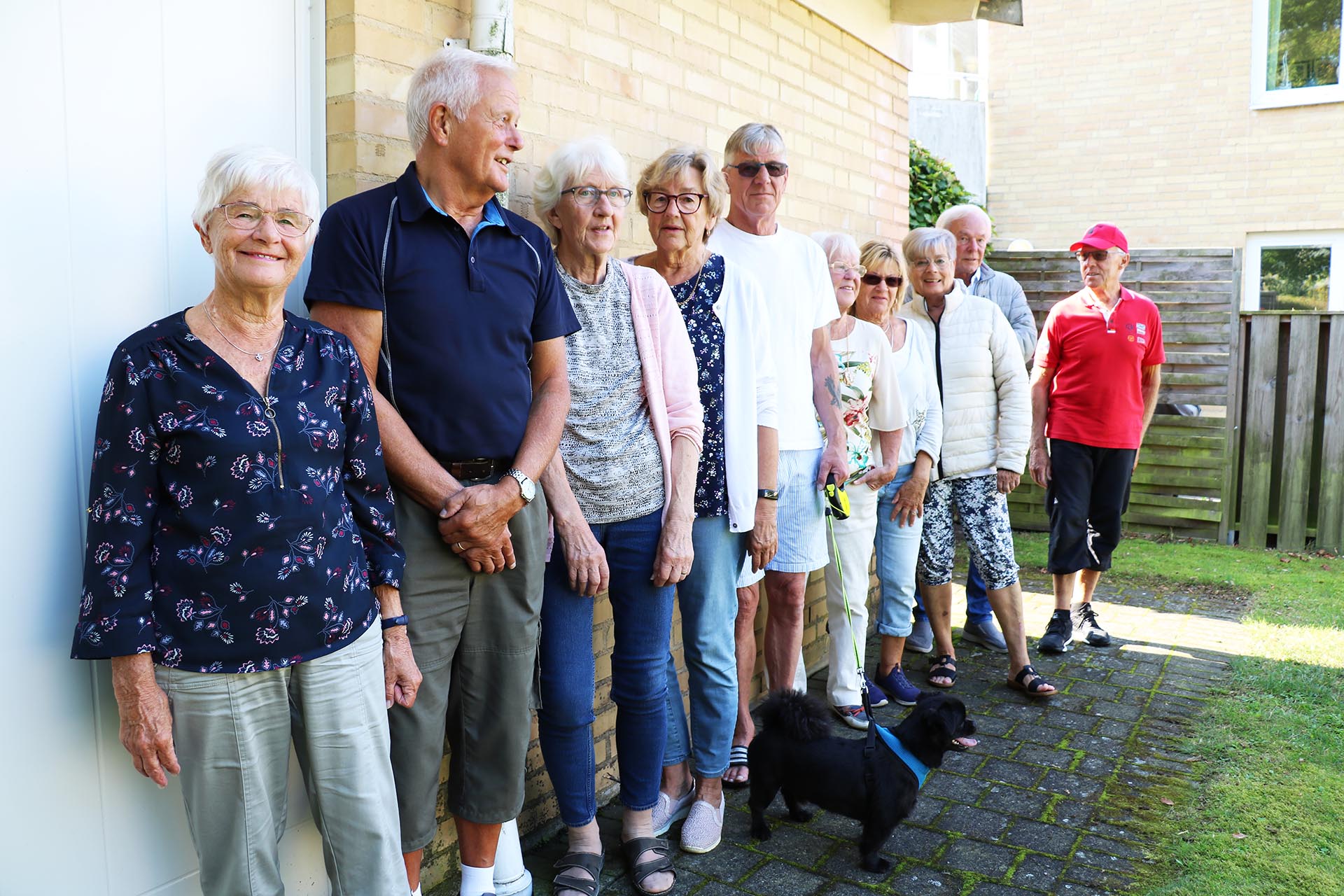 9 äldre personer står på rad längst en husvägg