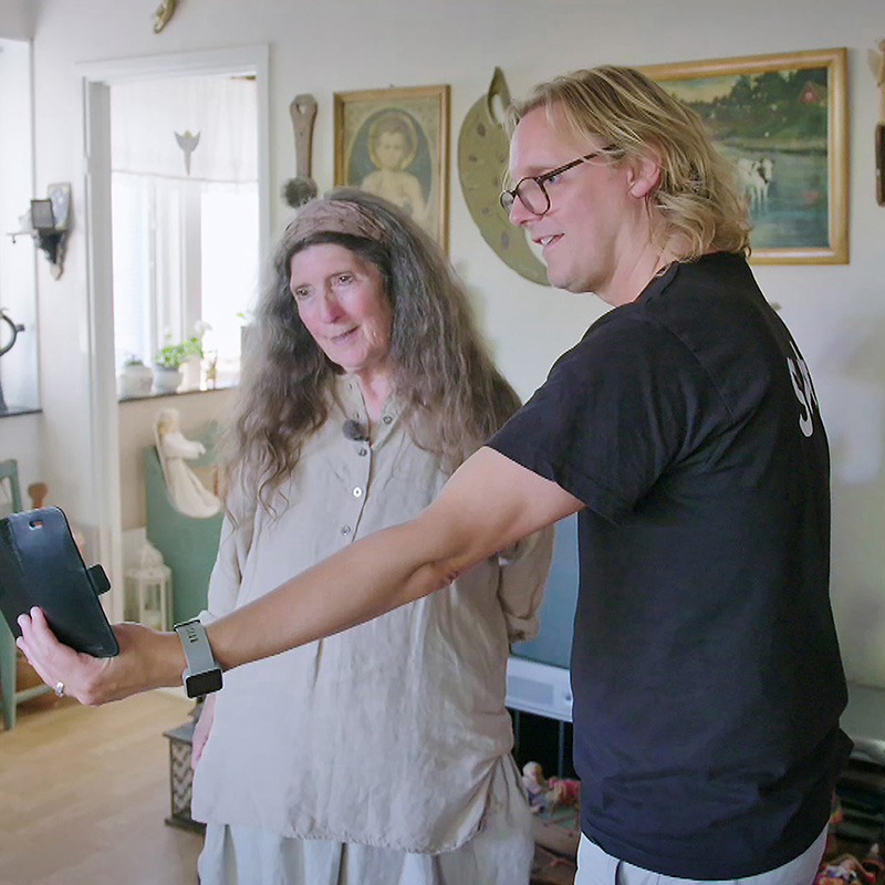 En kvinna med långt grått hår och en kille med en svart tröja står i ett rum och tittar på en mobil