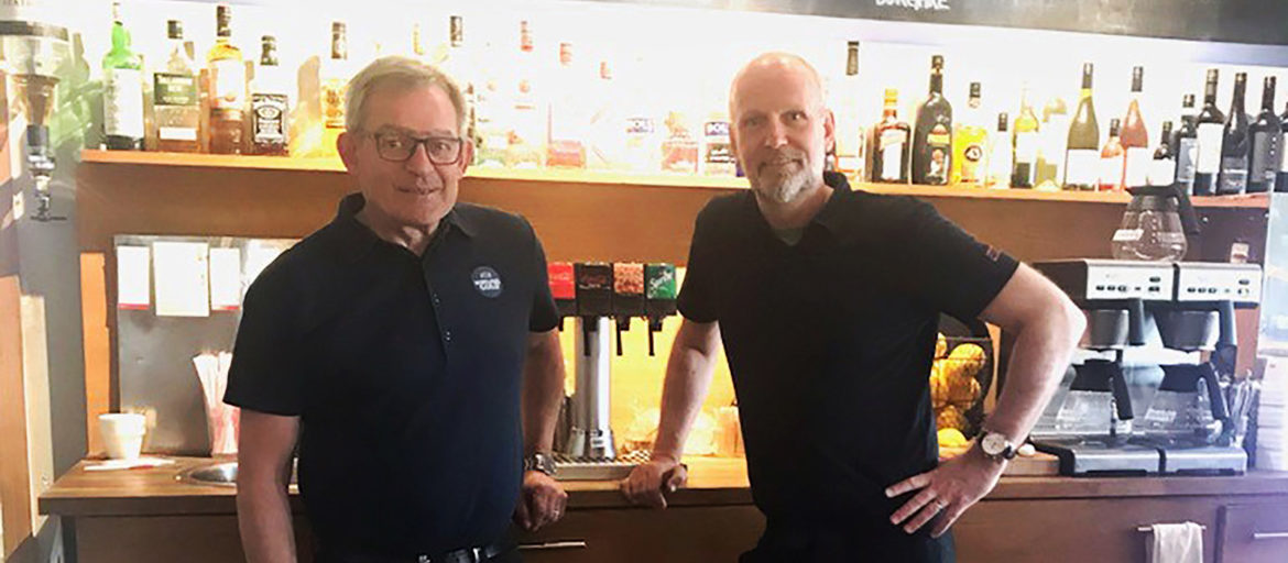 Två män i svarta pikétröjor står framför en bar