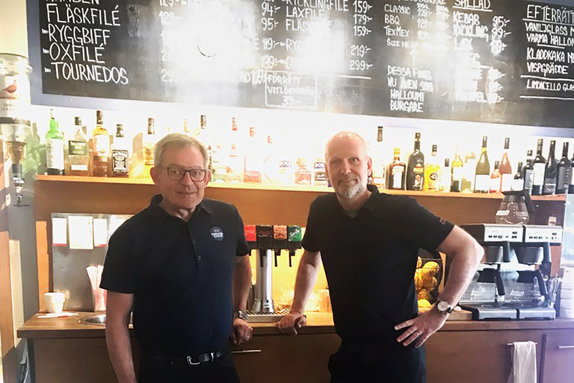 Två män i svarta pikétröjor står framför en bar