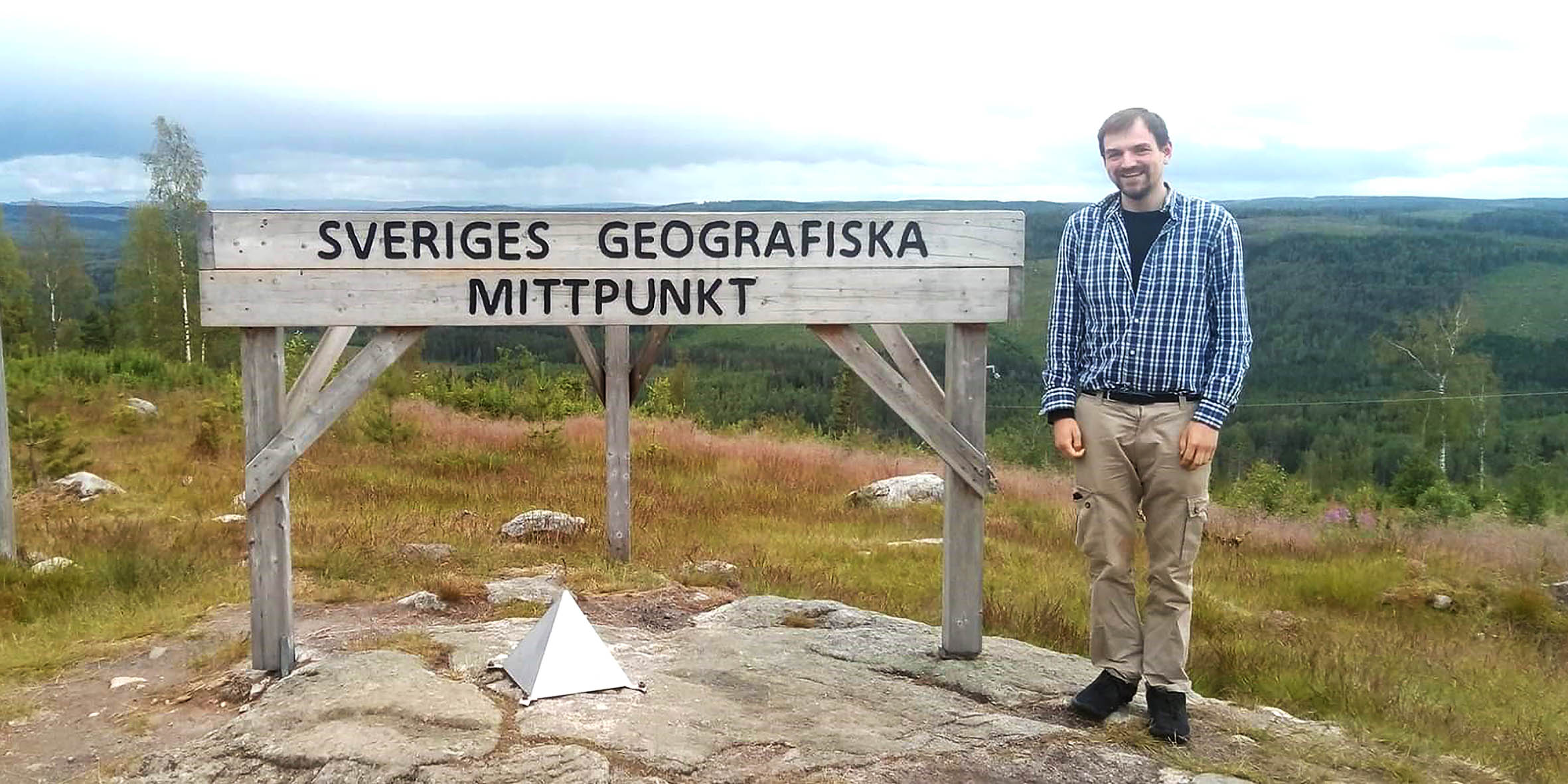En man står på ett berg framför en skylt som det står Sveriges geografiska mittpunkt på
